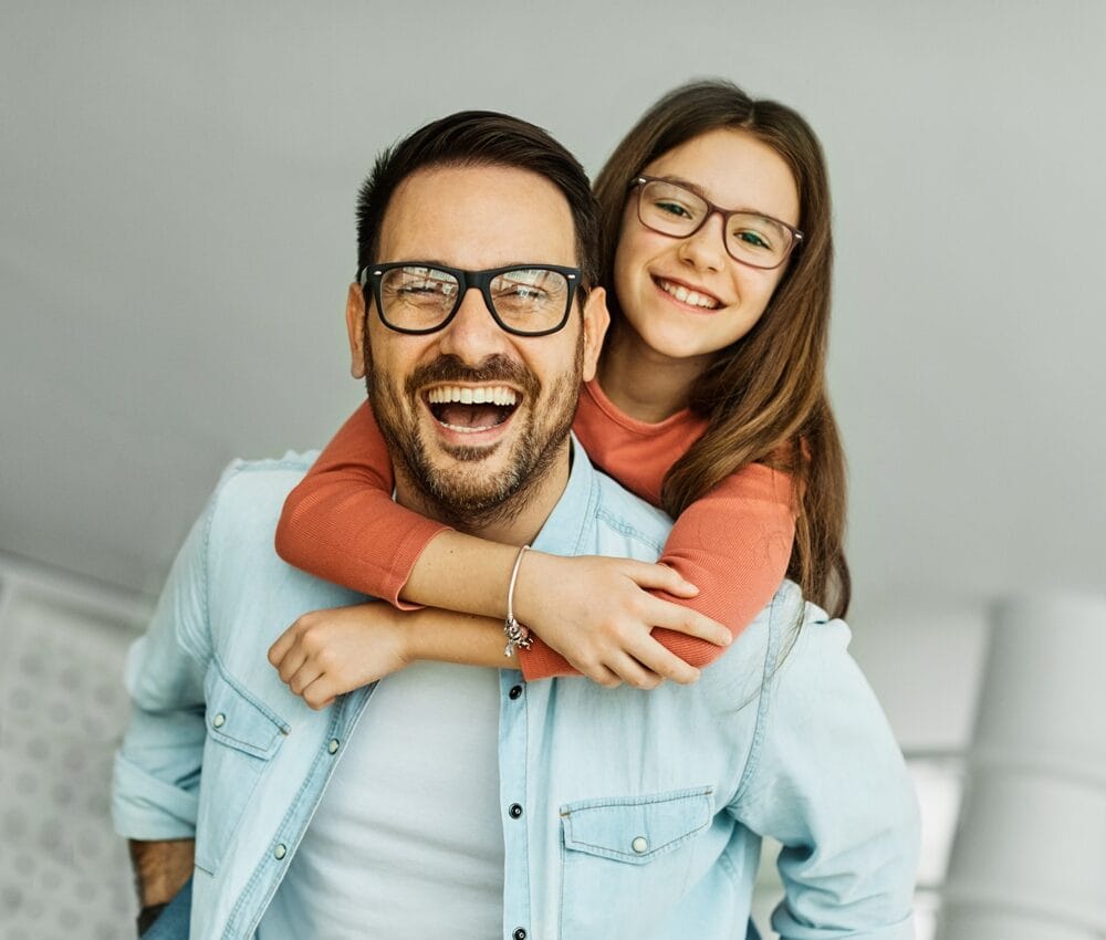 A cheerful father wearing glasses gives his smiling daughter, also in glasses, a piggyback ride as she hugs him from behind in a bright indoor setting. - Falcon Vision